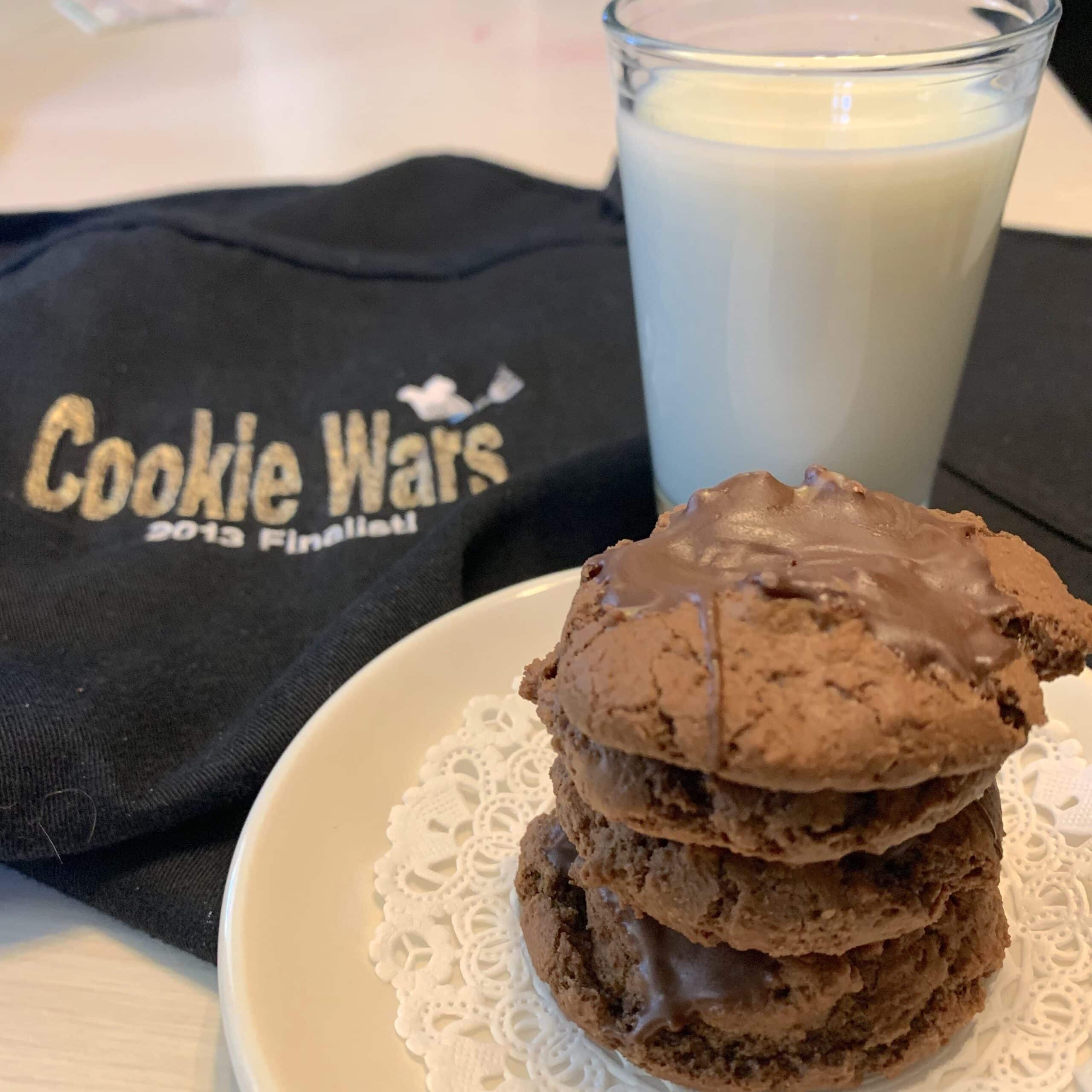 A stack of chocolate mint cookies stands on a plate next to a glass of cold, white milk. In the backrown is a black apron with "Cookie Wars" emgroidered on it