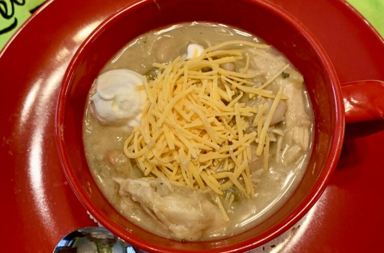 Creamy white chicken chili in a red mug placed o a red plate. greet background with partial words Lets Cook