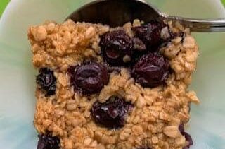 Close of of a square of baked oatmeal studded with blueberries in a white Depression Glass bowl and a spoon.