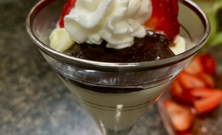 A cordials glass holds creamy panna cotta topped with chocolate ganache, whipped cream and strawberries